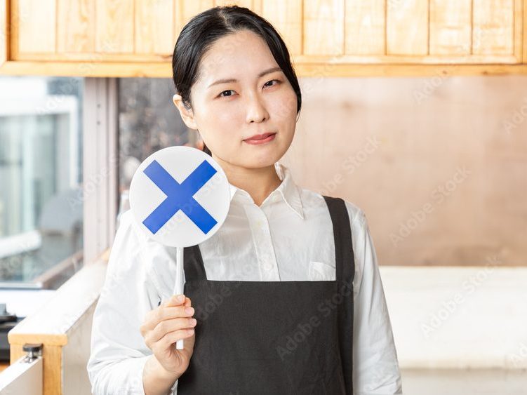 エプロンを付けたカフェ店員が×マークを手に持っている写真。就労移行支援中のアルバイトは原則禁止です。